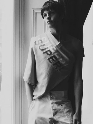 Young man wearing a graphic t-shirt with large text standing near a door frame in a black and white editorial photograph.