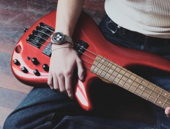 Close-up of a person holding a metallic red electric bass guitar while wearing dark jeans and a white ribbed sweater.