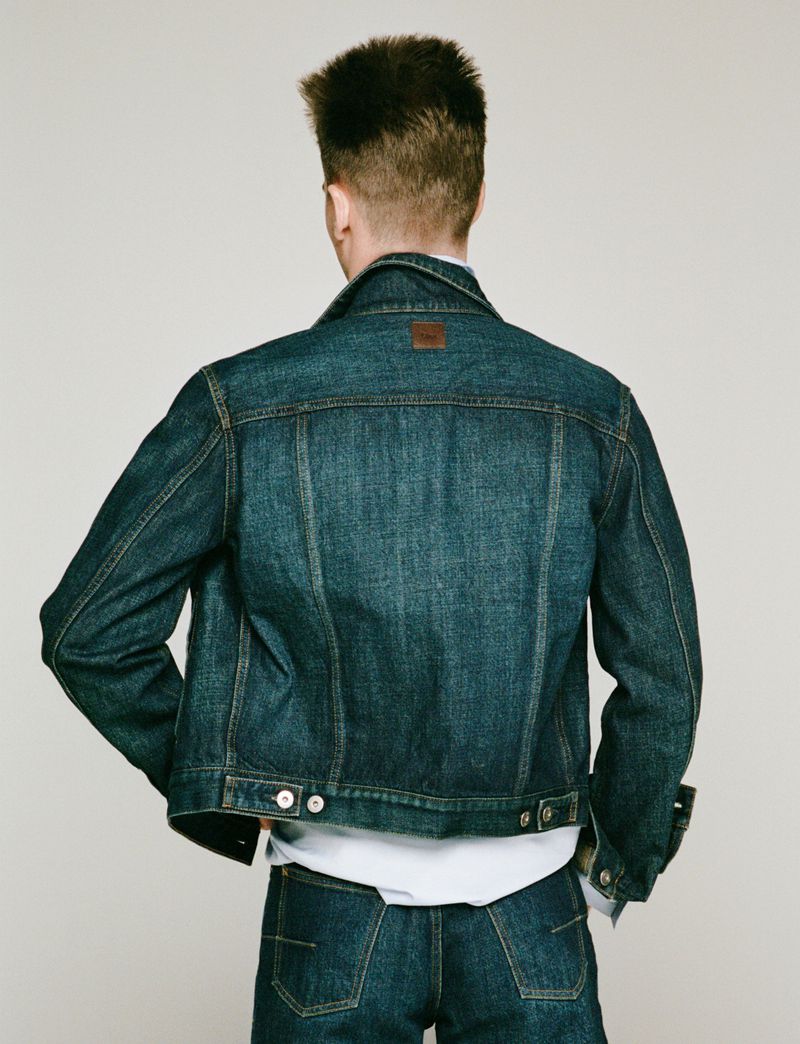 A man in a denim jacket and jeans stands against a neutral backdrop, showcasing the back view of the outfit in a studio setting.
