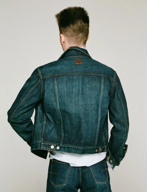 A man in a denim jacket and jeans stands against a neutral backdrop, showcasing the back view of the outfit in a studio setting.