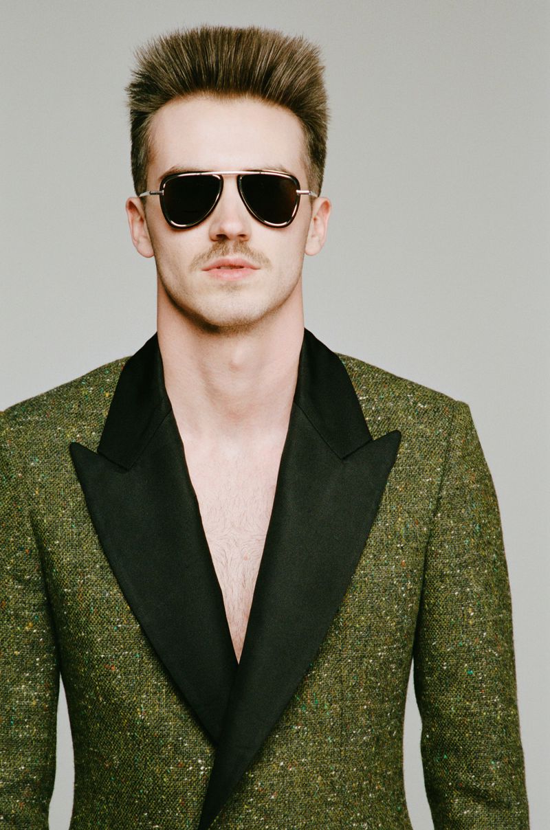 Portrait of adult man wearing aviator sunglasses and green tweed blazer with black lapels, posing indoors against a grey backdrop.