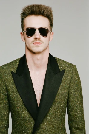 Portrait of adult man wearing aviator sunglasses and green tweed blazer with black lapels, posing indoors against a grey backdrop.