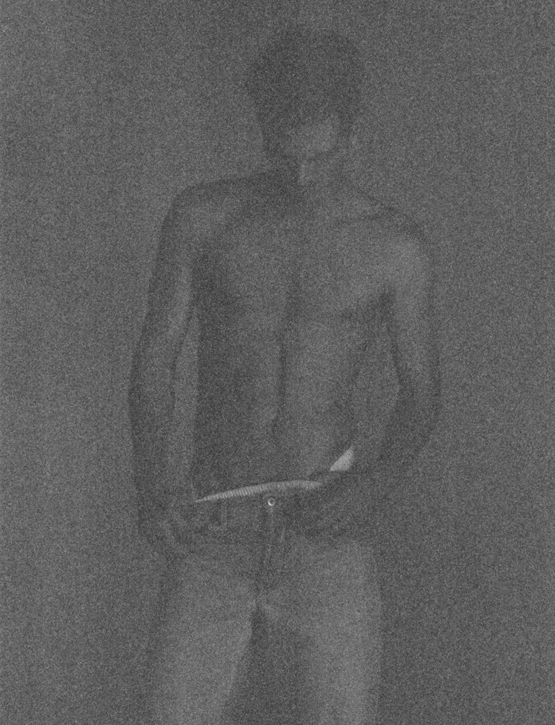 Monochromatic studio portrait of a muscular adult male, shirtless and wearing jeans, standing against a grey gradient background with soft lighting.