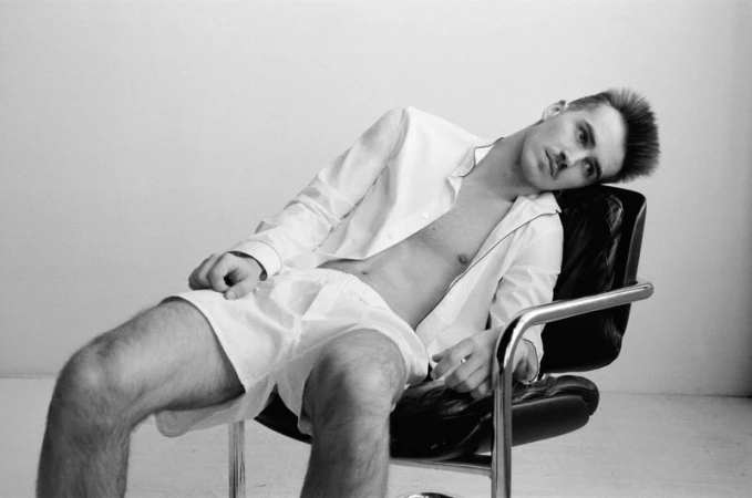 Black and white photo of a man in a shirt and boxer shorts reclining in a black chair against a white wall in a photo studio.