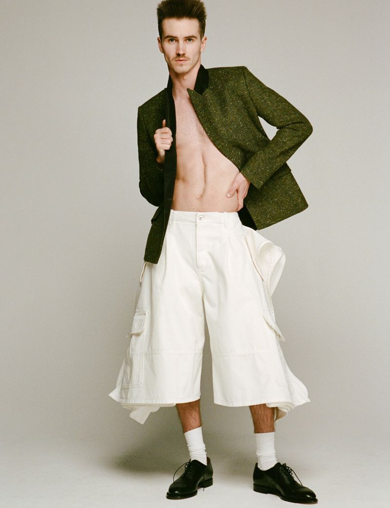 Male model in avant-garde fashion posing in a green jacket and oversized white culottes against a neutral studio background.