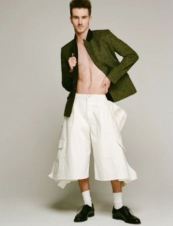 Male model in avant-garde fashion posing in a green jacket and oversized white culottes against a neutral studio background.