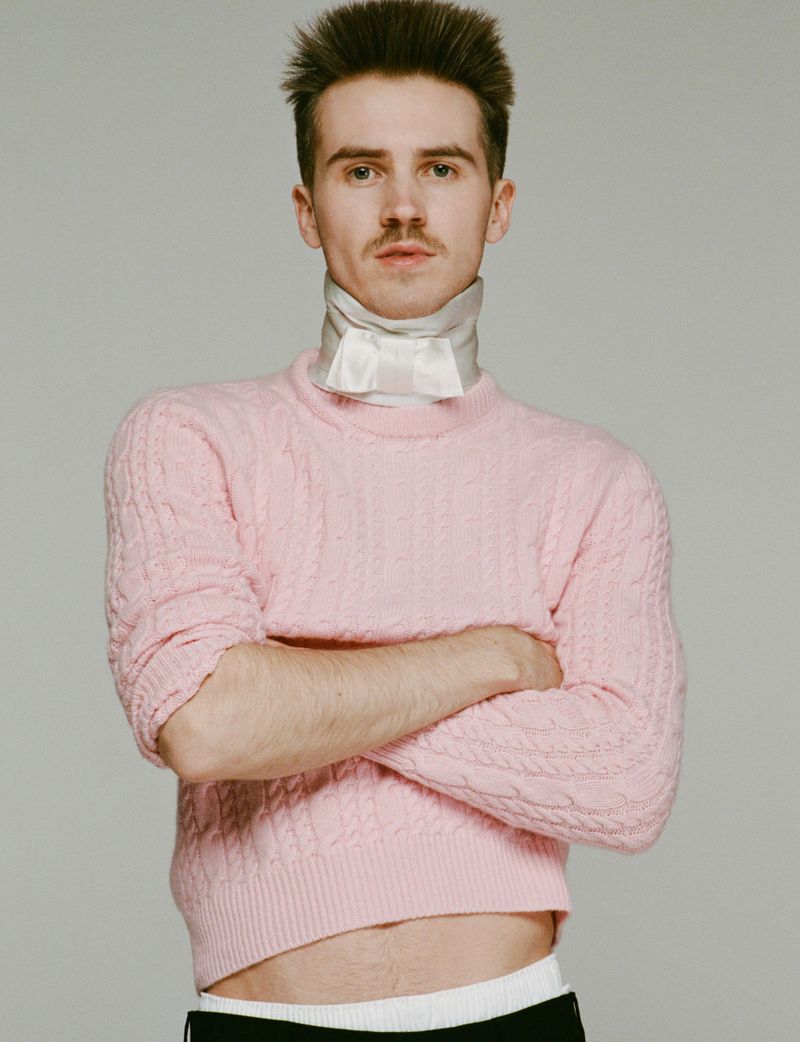 A portrait shot of a young adult male with a mustache, wearing a pink sweater and a white neck scarf, posing indoors against a grey background.