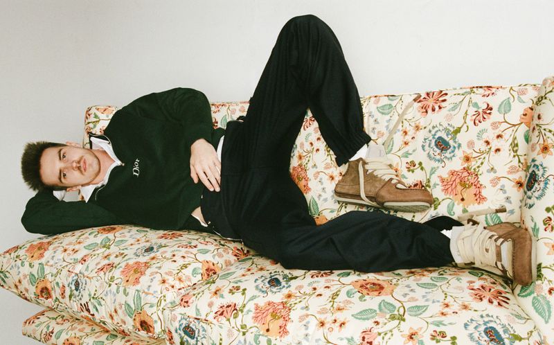 Adult male lying on a floral patterned sofa, wearing a dark green collared sweater, black pants, and brown high-top sneakers in an indoor setting.