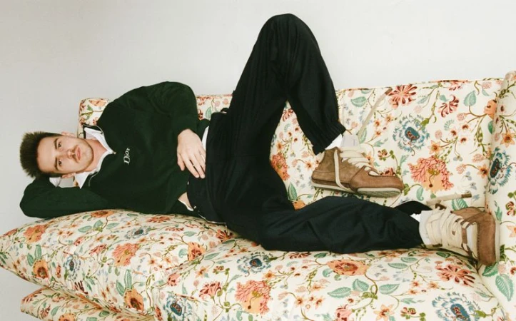 Adult male lying on a floral patterned sofa, wearing a dark green collared sweater, black pants, and brown high-top sneakers in an indoor setting.