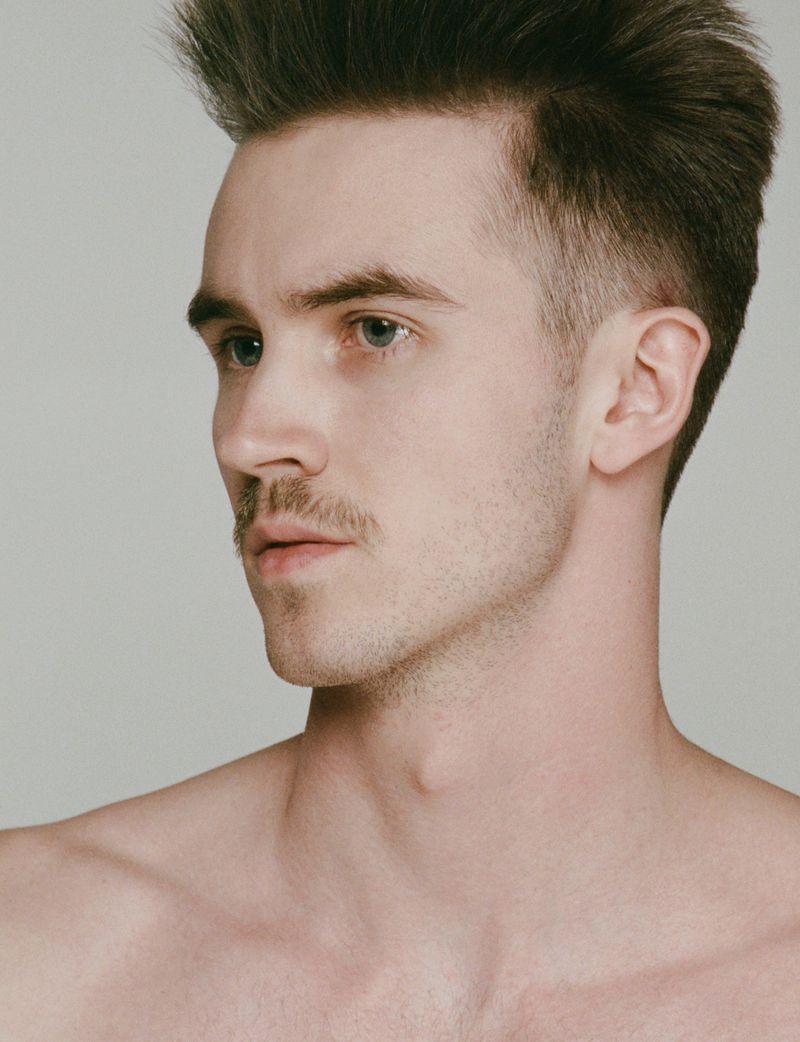 Close-up headshot of a Caucasian adult male with short brown hair, a mustache, and bare shoulders against a neutral grey background.