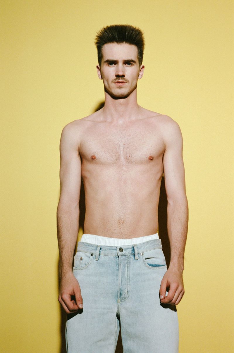 A shirtless man with a mustache stands against a yellow backdrop wearing light-wash jeans. The lighting casts a subtle shadow.