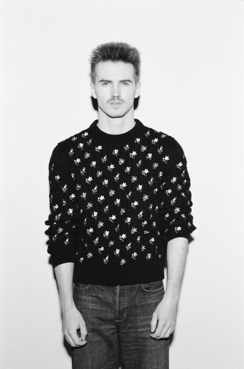 Portrait of adult man with distinct hairstyle and mustache wearing a patterned black sweater and denim jeans against white backdrop.