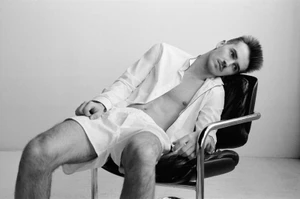 Monochrome portrait of a young man reclining in a modern leather and metal chair wearing an unbuttoned white shirt and shorts.