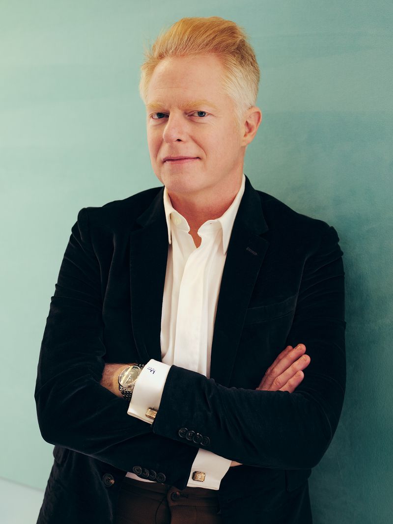 Headshot of an adult male with fair skin and reddish blond hair, wearing a black velvet blazer, white collared shirt, and brown pants, arms crossed.
