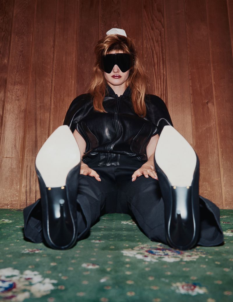 Stylish woman with visor and black corset poses seated on green floral carpet in front of wood paneled wall, showcasing her heels.