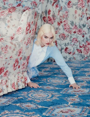 A woman in a blue and white shirt crawls on a blue patterned carpet in a room with a floral print couch and wallpaper.