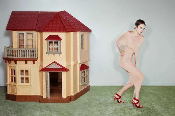 Fashion shot of a woman in a nude dress and red heels, posing next to a dollhouse with a red roof and light-yellow siding, indoors.