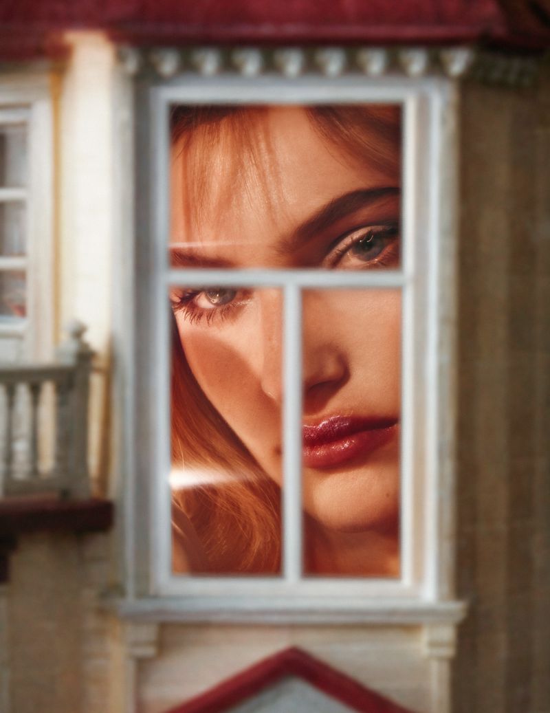 Close-up of a woman's face seen through a window with white frames and blurred exterior architectural details of a building.