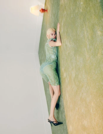 Adult female in a patterned short dress and heels lies on a green carpet next to a white wall. A lamp illuminates the scene.