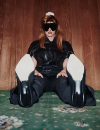 Model seated on a patterned green carpet wearing black clothing and large sunglasses with wooden paneling behind