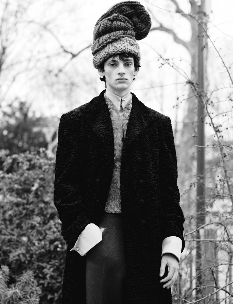 Black and white portrait of a slender male model in a textured coat and oversized knit hat, standing outdoors against a blurred background.