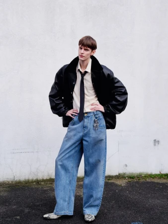 Stylish young adult in an oversized black bomber jacket, light shirt, tie, and wide-leg jeans, posing against a textured white wall outdoors.