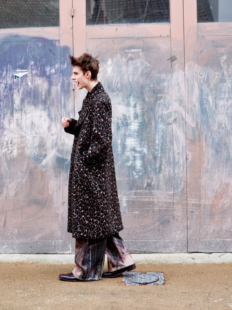 Side view of a young adult male with dark hair, wearing a patterned coat and wide-leg pants, standing in front of an aged door. 