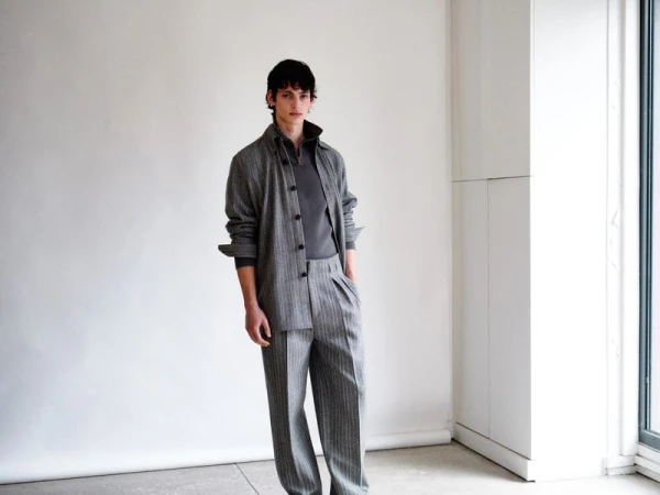 A young adult male poses indoors wearing a gray button-down shirt, t-shirt, and striped dress pants in front of a white wall and window.