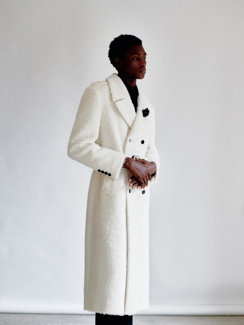 Side view of an adult Black male wearing a double-breasted white overcoat in a minimalist studio, looking off to the right.
