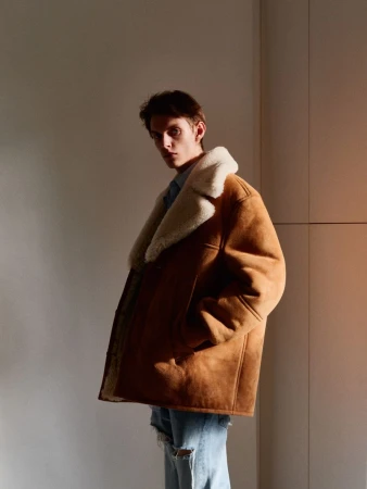 Man posing in shearling-lined jacket and ripped jeans in a studio with soft lighting. Fashion editorial with neutral background.