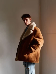Young man wearing a brown shearling coat and distressed jeans standing indoors against a plain wall.