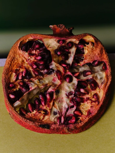 Halved pomegranate with ruby seeds and thick rind sits on a pale green surface against a dark green background.