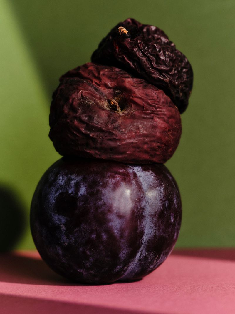Close-up studio shot of a stack of plums with varying degrees of ripeness, set against a vibrant green and pink backdrop.