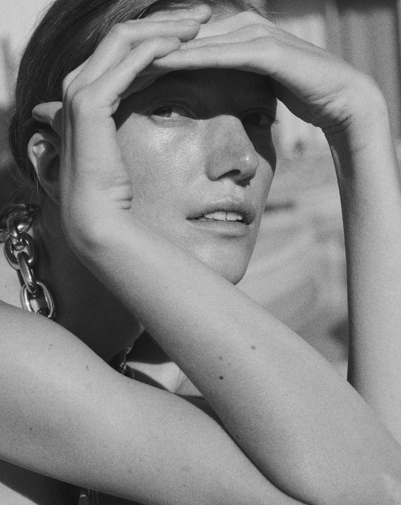Close-up black-and-white portrait of a fair-skinned woman shielding her eyes with her hands while wearing a chain necklace.