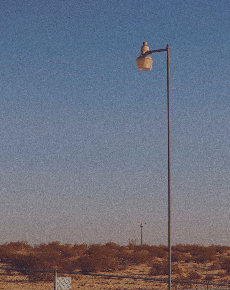 A tall metal street light in a desert setting under a clear sky. The light's design is vintage, casting a warm glow on the arid landscape.