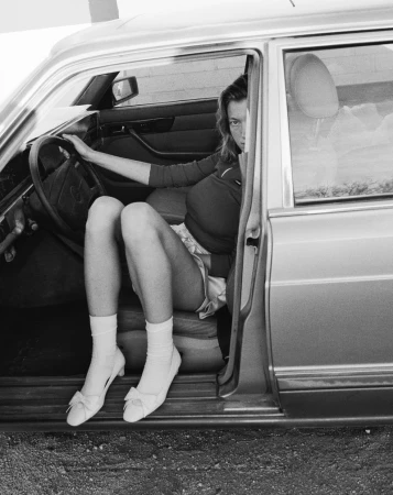 Black and white photo of a woman with fair skin sitting in the driver's seat of a vintage car, wearing white shoes and socks.