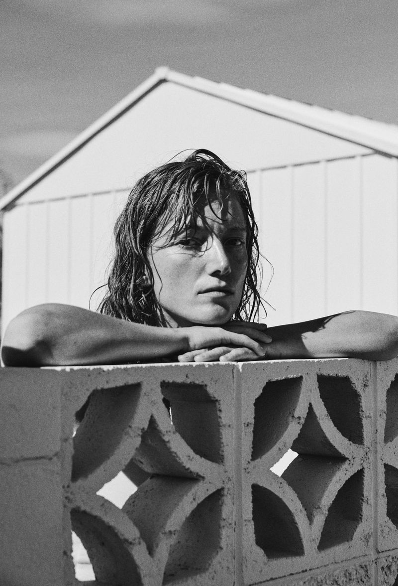 Monochrome portrait of a young adult leaning on a concrete block wall, with a white shed and sky visible in the background.