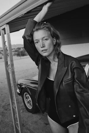 Woman in a swimsuit and jacket poses next to a classic car, under a carport in an outdoor setting with a desert landscape in the distance.