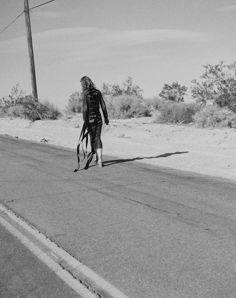 A slender adult poses on a desert road in a dark outfit, ribbons trailing behind, against a backdrop of arid vegetation and clear sky.
