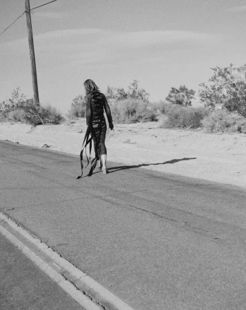 A slender adult poses on a desert road in a dark outfit, ribbons trailing behind, against a backdrop of arid vegetation and clear sky.