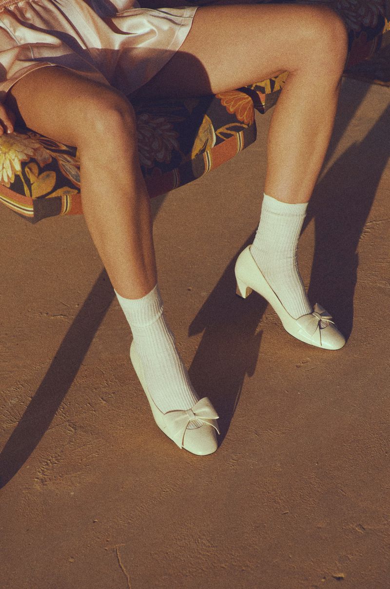 Woman's legs on a floral cushion chair, wearing white socks and white high heel shoes with bow, casting shadows on a tan surface.