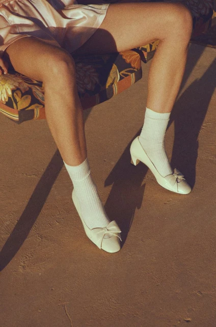 Woman's legs on a floral cushion chair, wearing white socks and white high heel shoes with bow, casting shadows on a tan surface.