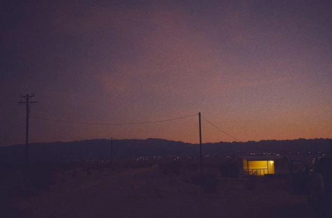 Twilight landscape featuring a utility pole, small building with illuminated windows, and a mountain range under a gradient sky.