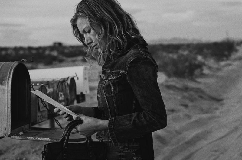 Black and white photo of woman in denim jacket reading letter at rural mailbox in desert landscape, overcast sky, film photography.