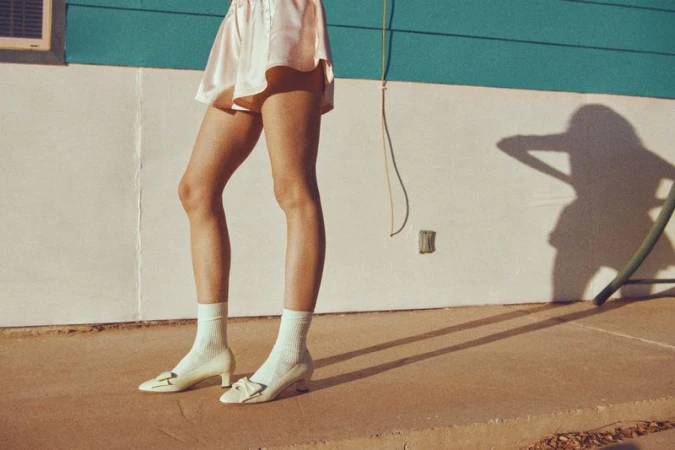 A woman's lower body is displayed, featuring a pink skirt, white socks, and light yellow heeled shoes in an outdoor setting.