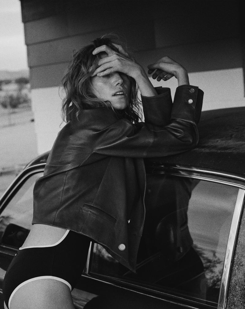 Black and white shot of a woman in a leather jacket posing against a car, with a desert landscape and building in the background.