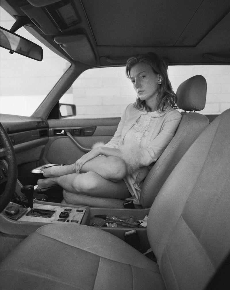 Black and white portrait of a young woman seated inside a car, wearing a light-colored dress and cardigan, with light streaming in.