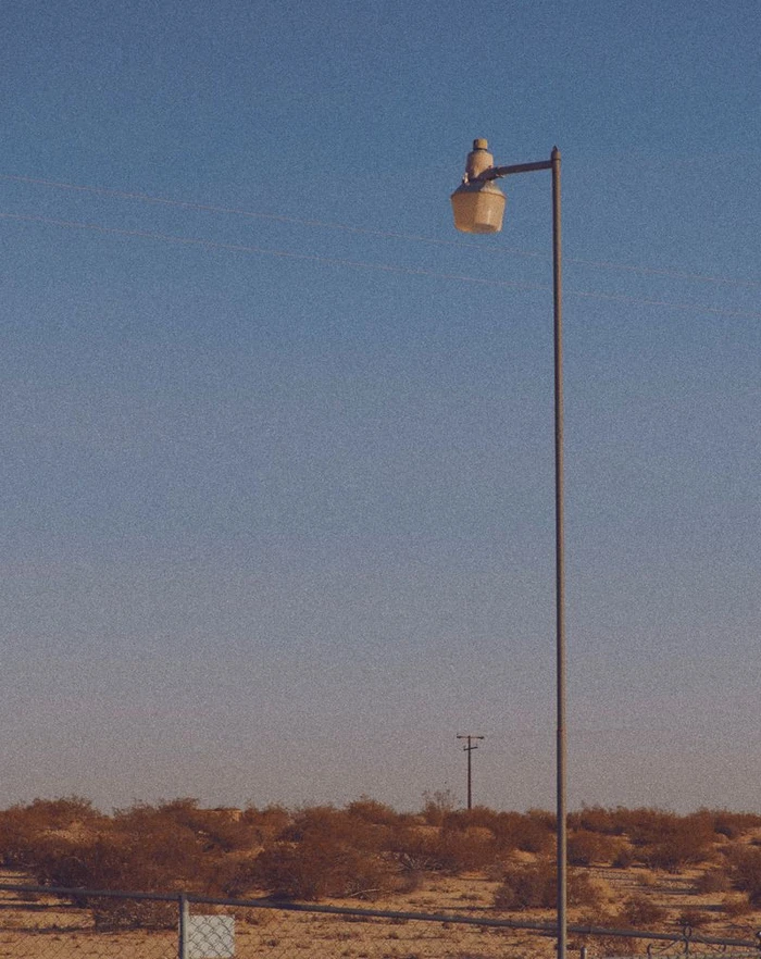 A lone streetlamp standing against a dusk sky over a desert landscape with sparse low shrubs and a wire fence.