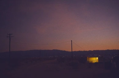 Dusk landscape in the desert with utility poles, distant mountains, and a single illuminated cabin under a purple sky