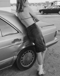 Model leaning against a parked car wearing a skirt and socks, captured in monochrome.